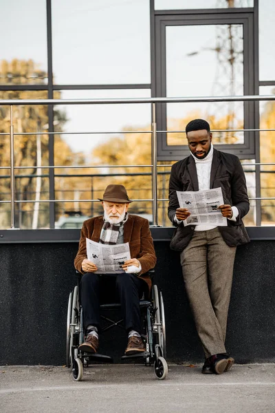 Sénior discapacitado hombre en silla de ruedas y guapo afroamericano hombre leyendo periódicos juntos en la calle - foto de stock