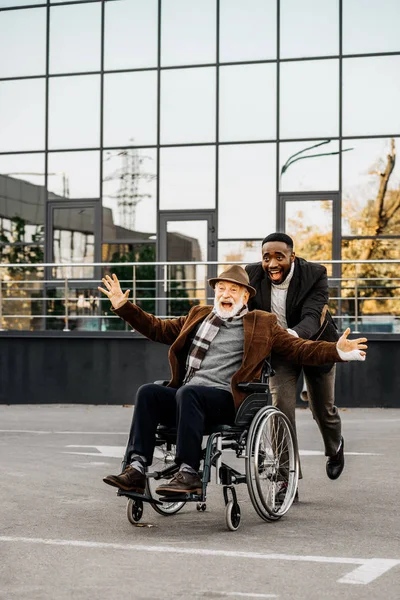 Laughing senior disabled man in wheelchair and african american man having fun while riding by street — Stock Photo