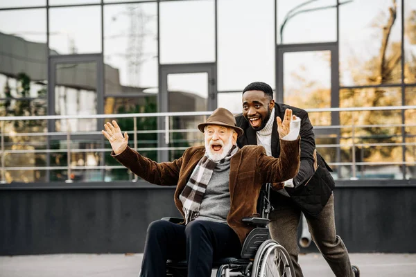 Happy senior disabled man in wheelchair and african american man having fun while riding by street — Stock Photo