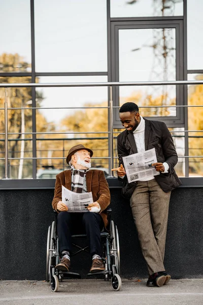 Anciano discapacitado en silla de ruedas y afroamericano leyendo periódicos juntos en la calle - foto de stock
