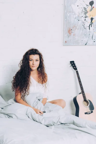 Hermosa joven sentada en la cama cerca de la guitarra acústica en casa - foto de stock