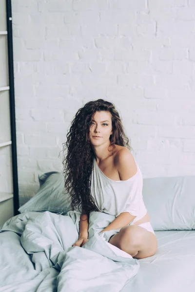 Attraktive junge Frau sitzt im Bett und schaut morgens zu Hause in die Kamera — Stockfoto