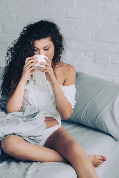 Приваблива кучерява жінка п'є каву в ліжку в ранковий час вдома — стокове фото