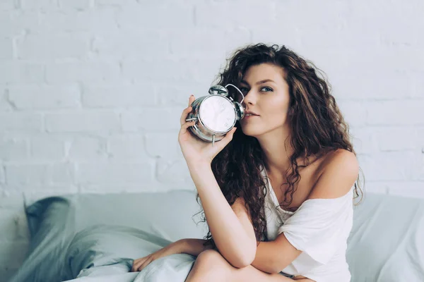 Cheerful curly young woman sitting on bed with alarm clock at home — Stock Photo