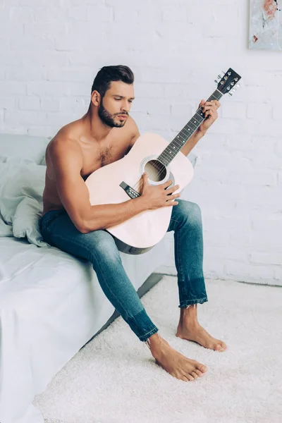 Homem sem camisa muscular em jeans tocando na guitarra acústica durante o horário da manhã na cama em casa — Fotografia de Stock