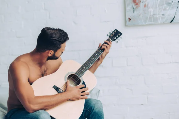 Jovem sem camisa homem em jeans tocando na guitarra acústica durante o horário da manhã na cama em casa — Fotografia de Stock