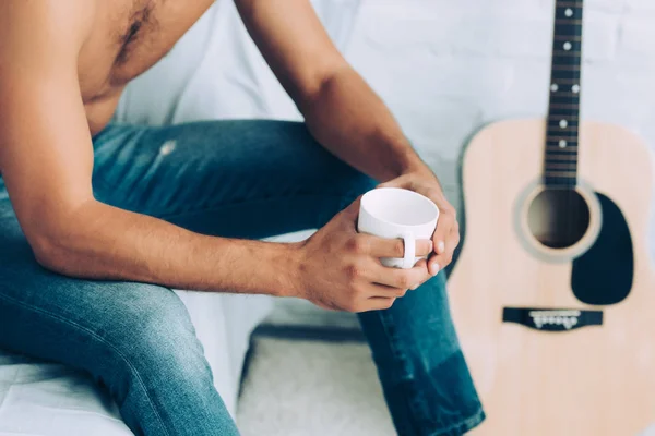 Частичный вид человека без рубашки в джинсах, пьющего чашку кофе в утреннее время в спальне дома — стоковое фото