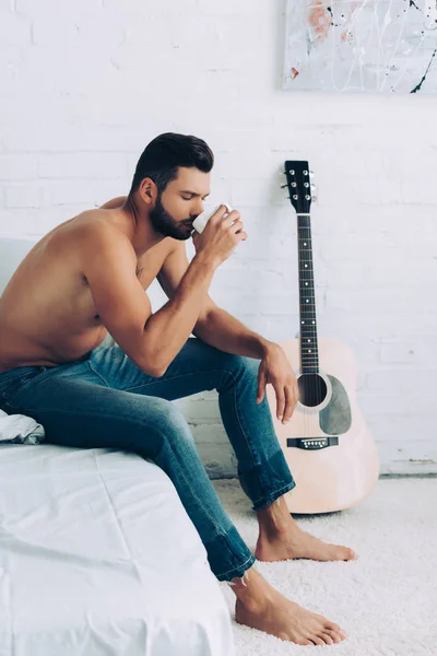 Shirtless muscular young man in jeans drinking coffee cup during morning time in bedroom at home — Stock Photo