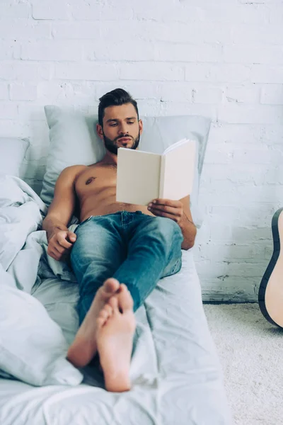 Сфокусированный без рубашки мускулистый мужчина читает книгу утром в спальне дома — стоковое фото