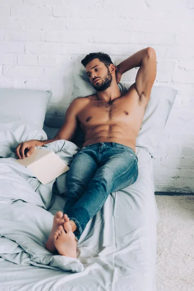 Junger muskulöser Mann ohne Hemd sitzt mit Buch zu Hause auf dem Bett — Stockfoto