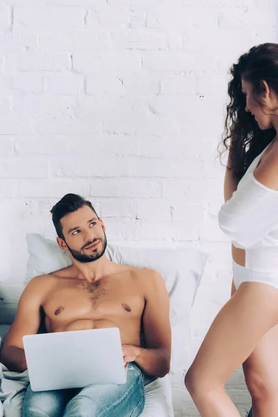 Smiling shirtless male freelancer with laptop sitting on bed and talking to seductive girlfriend at home — Stock Photo