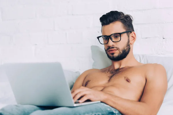 Freelancer masculino sem camisa muscular em óculos trabalhando no laptop na cama em casa — Fotografia de Stock