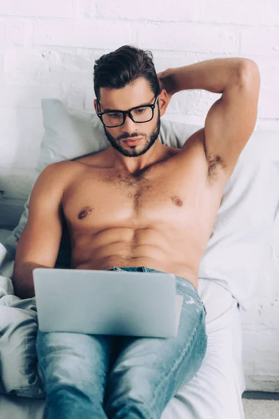 Vue grand angle de torse nu musclé mâle pigiste dans les lunettes de travail sur ordinateur portable dans le lit à la maison — Photo de stock