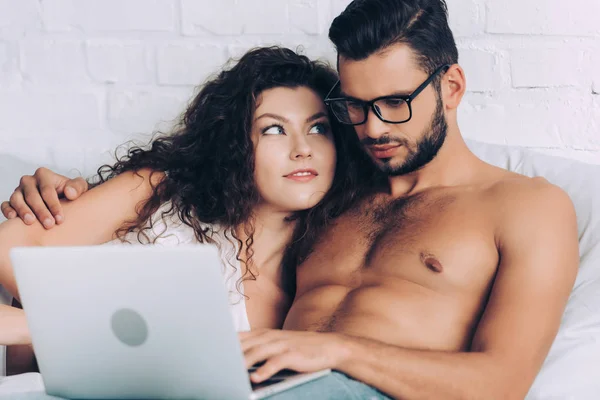 Junge hemdlose männliche Freiberufler mit Brille arbeiten am Laptop und umarmen Freundin im Schlafzimmer zu Hause — Stockfoto