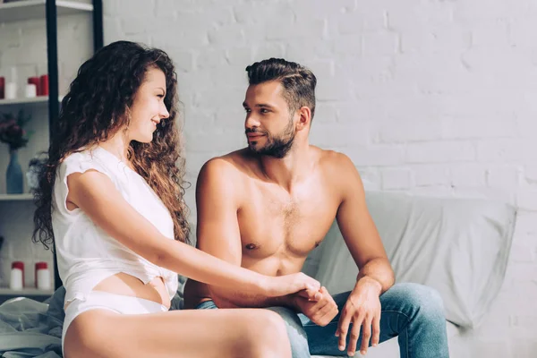 Cheerful sexy girl in white underpants holding hand of boyfriend in bedroom at home — Stock Photo