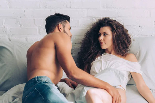 Handsome muscular man in jeans and his curly attractive girlfriend on bed at home — Stock Photo