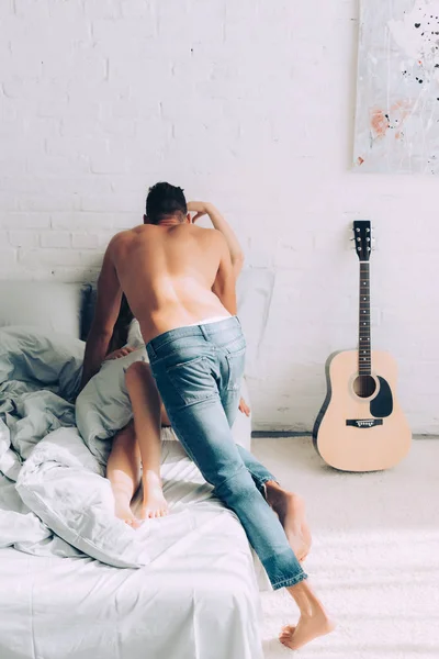 Vue arrière de l'homme musclé torse nu en jeans avec petite amie sur le lit à la maison — Photo de stock