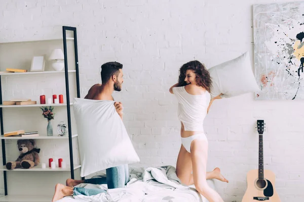 Happy young couple having fun and fighting by pillows in bedroom at home — Stock Photo