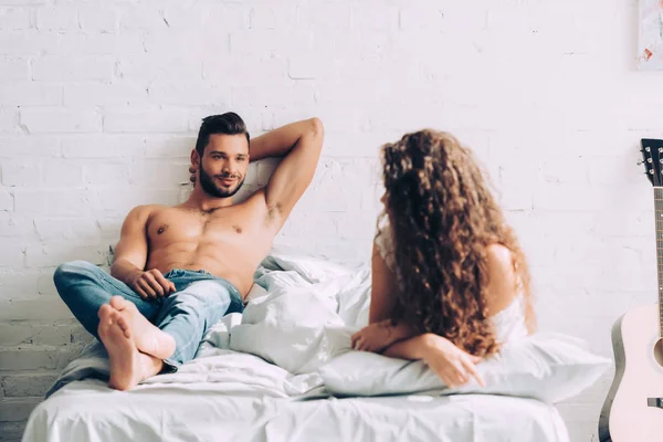 Muskulöser, hemdloser Mann in Jeans, der morgens zu Hause im Schlafzimmer mit seiner lockigen Freundin spricht — Stockfoto