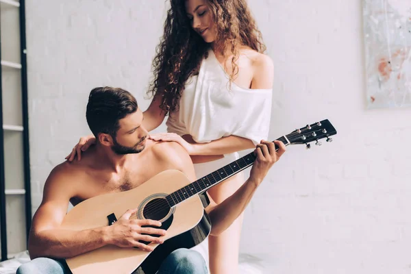 Bel homme torse nu jouant sur la guitare acoustique tandis que sa petite amie debout près dans la chambre à coucher à la maison — Photo de stock
