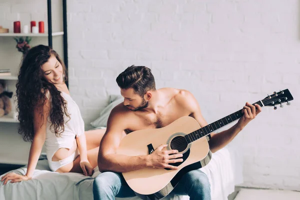 Musculoso hombre sin camisa tocando la guitarra acústica mientras su novia se sienta cerca en el dormitorio en casa - foto de stock