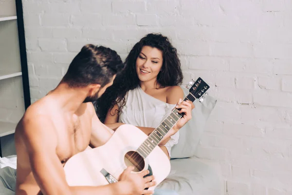 Homme jouant sur la guitare acoustique tandis que sa petite amie heureuse assis près dans la chambre à coucher à la maison — Photo de stock