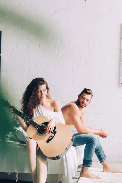 Selective focus of curly woman playing on acoustic guitar while her boyfriend sitting near on bed at home — Stock Photo