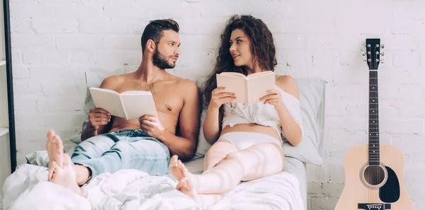 Feliz joven pareja sentada con libros y mirándose en la cama en casa - foto de stock