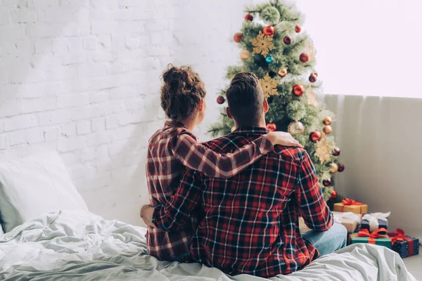Visão traseira do casal sentado na cama e olhando para a árvore de natal decorada em casa — Fotografia de Stock