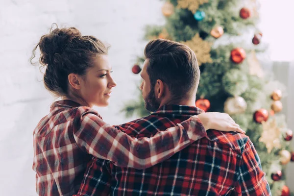 Casal em camisas xadrez olhando uns para os outros perto da árvore de natal em casa — Fotografia de Stock