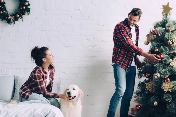Vue latérale de fille caressant golden retriever tandis que son petit ami décorer arbre de Noël à la maison — Photo de stock