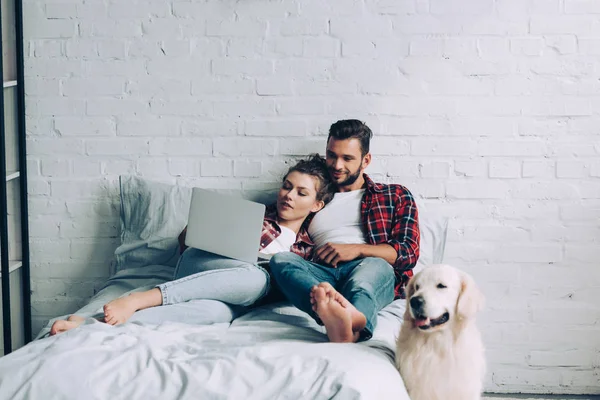 Jovem casal deitado na cama e observando laptop enquanto seu golden retriever de pé perto de casa — Fotografia de Stock