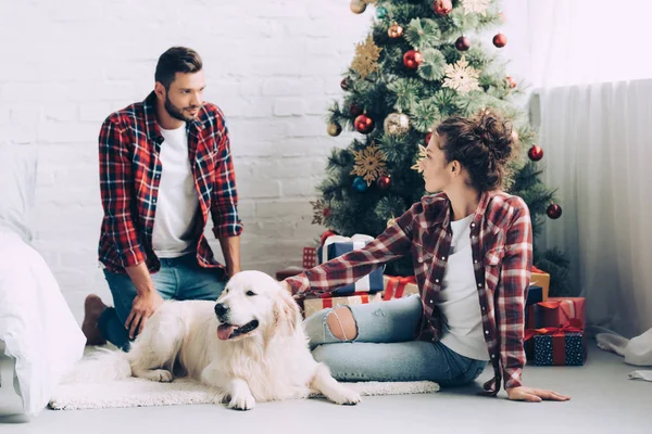 Side view of beautiful girl petting dog and talking to boyfriend near christmas tree at home — Stock Photo