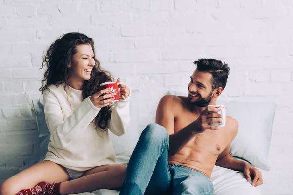 Rindo jovem casal com xícaras de café sentado na cama durante a hora da manhã em casa — Fotografia de Stock