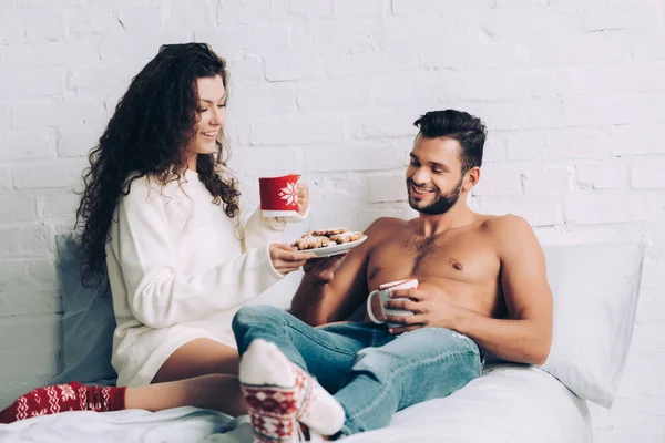 Smiling young woman holding plate with cookies while her shirtless boyfriend sitting with coffee cup in bedroom at home — Stock Photo