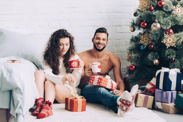 Glückliches junges Paar mit Kaffeetassen in der Nähe von Geschenkboxen und Weihnachtsbaum zu Hause — Stockfoto