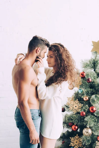 Alegre joven mujer abrazando sin camisa novio cerca de árbol de Navidad en casa - foto de stock
