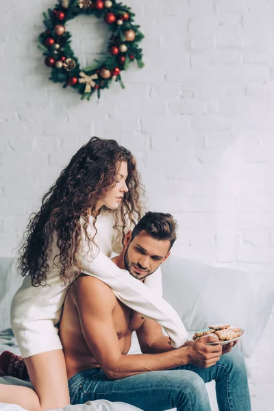 Pretty curly girl hugging shirtless boyfriend while he holding plate with ginger cookies in bedroom at home — Stock Photo