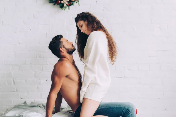 Vista lateral de la mujer rizada sexy sentado en el novio sin camisa en la cama en casa — Stock Photo