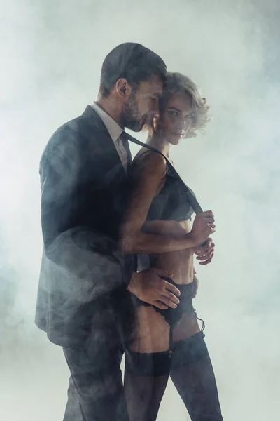 Мужчина обнимает блондинку замечательную женщину в нижнем белье, пока она тянет его за галстук на сером фоне дыма — стоковое фото