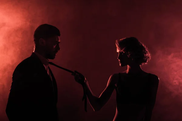 Чувственная женщина тянет мужчину за галстуком на красном фоне дыма — стоковое фото