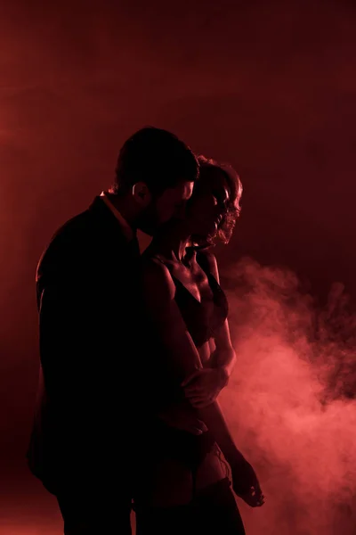 Man tenderly hugging beautiful woman and kissing her in neck on red smoke background — Stock Photo