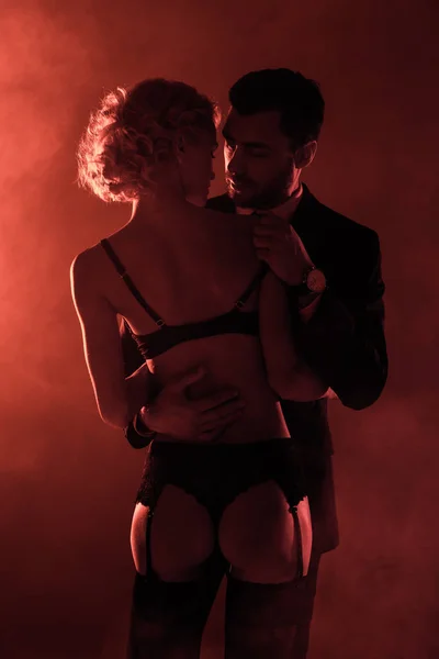 Vue arrière de la femme séduisante en lingerie regardant l'homme en costume sur fond de fumée rouge — Photo de stock