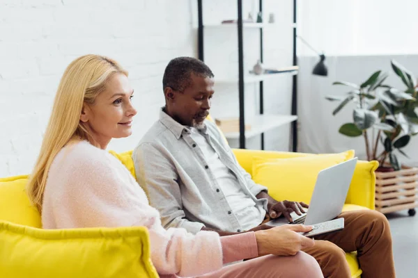 Maturo uomo africano americano utilizzando il computer portatile mentre la donna seduta sul divano con telecomando — Foto stock