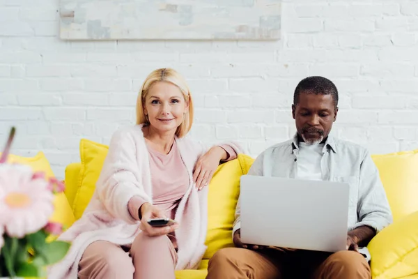 Homme américain africain mature utilisant un ordinateur portable tandis que la femme assise sur le canapé avec télécommande — Photo de stock