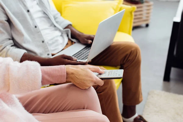Крупним планом афроамериканський чоловік використовує ноутбук, а жінка натискає кнопки на пульт дистанційного керування — стокове фото