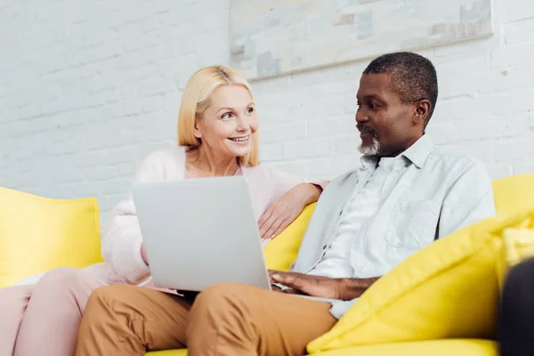 Mujer feliz sentada en el sofá con el hombre afroamericano y el uso de la computadora portátil - foto de stock