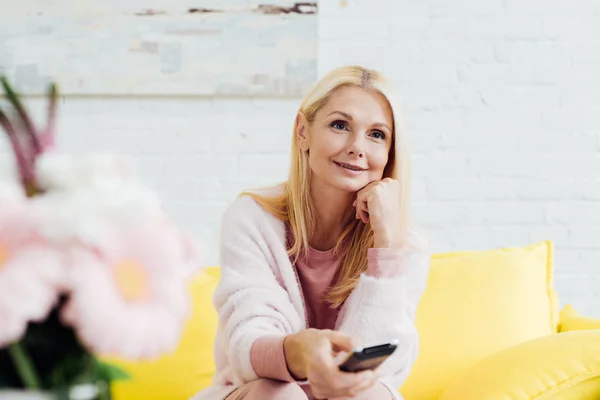 Belle femme mûre souriante assise sur un canapé jaune vif avec télécommande tv — Photo de stock