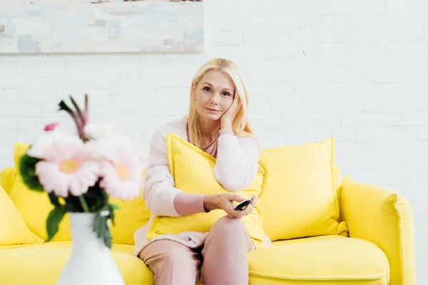 Attractive mature woman sitting on bright yellow sofa with tv remote controller — Stock Photo