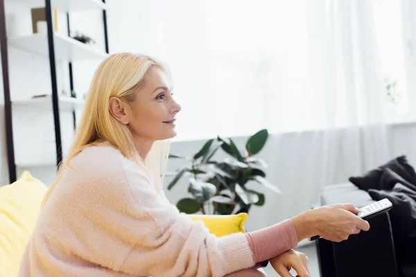 Beautiful smiling mature woman sitting on bright yellow sofa and pushing buttons on tv remote controller — Stock Photo